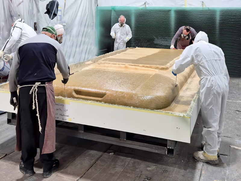 Auf dem Bild sind mehrere Personen in Schutzkleidung zu sehen, die an der Herstellung oder Bearbeitung eines großen Formstücks arbeiten.