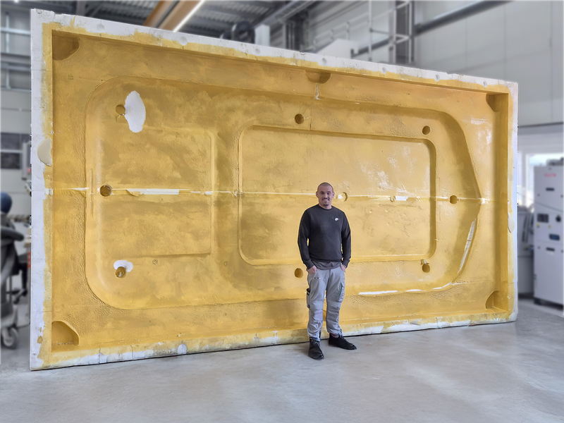 Auf dem Bild steht eine Person vor einer großen, gelben Form, wahrscheinlich für die Herstellung von Kunststoffteilen, in einer Werkstatt oder Fabrik.