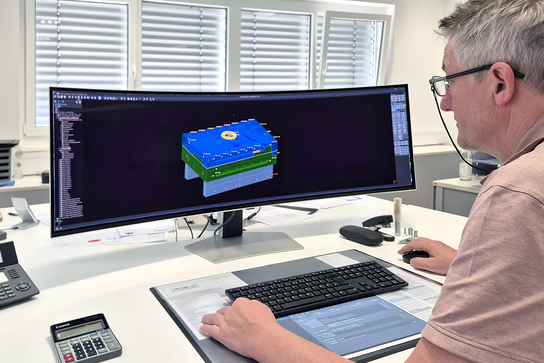 Auf dem Bild ist ein Mann zu sehen, der an einem Computer mit einem großen Bildschirm arbeitet, auf dem ein 3D-Modell angezeigt wird.