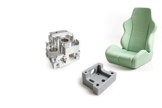 Auf dem Bild sind verschiedene Teile zu sehen: ein bearbeitetes Metallteil, eine graue Kunststoffform und ein gepolsterter Sitz.