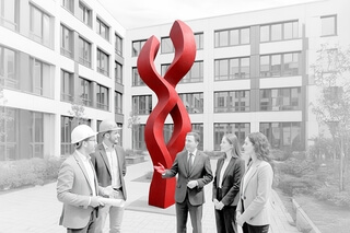 Auf dem Bild ist eine Gruppe von fünf Personen in einem Innenhof zu sehen, die vor einer großen roten Skulptur stehen und miteinander sprechen.