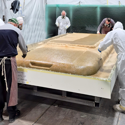 Auf dem Bild sind mehrere Personen in Schutzkleidung zu sehen, die an der Herstellung eines großen Kunststoffteils arbeiten.